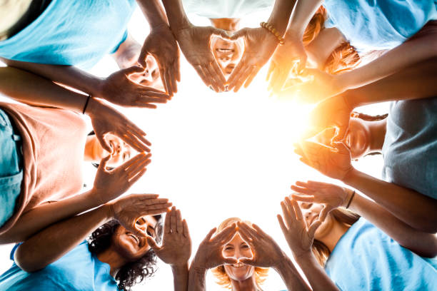 Multiracial group of diverse people stand in circle as community volunteers to show support and commitment to teamwork success togetherness making hand gesture in concept symbol sign of heart shape outdoors with sky  and sunlight stock photo