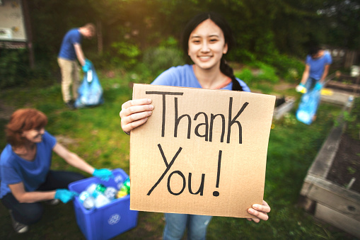 Young Asian woman holds thank you sign for gratitude in community garden park while diverse multiracial group of people help to cleanup trash and recycle plastic bottles for environmental conservation volunteer