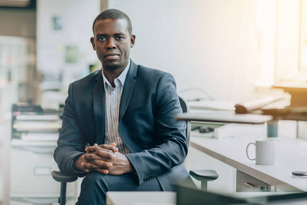 Portrait of Black mid adult businessman student sitting serious with hands clasped in bright business office wearing suit stock photo
