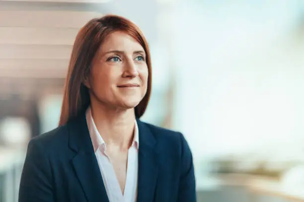 Photo of Portrait of Caucasian redhead mature businesswoman student smiling contented emotion satisfaction aspiration looking away forward in bright business office wearing suit businesswear