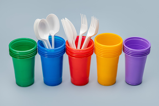 A set, in a row, of multi-colored disposable cups for drinks and cutlery. Grey background.