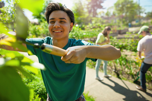Middle Eastern multiracial young man student smiles with pride and helps to trim bushes together with youth organization volunteer charity in local community garden public park in summer