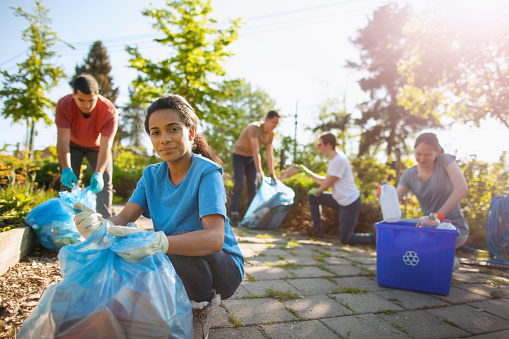 Portrait of Hispanic young woman student helping cleanup and recycle paper and plastic bottles within community garden public park with multiracial group of youth organization volunteer charity in summer