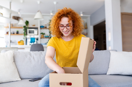 Beautiful young cheerful red hair woman unboxing a package in the living room of a new sweater.