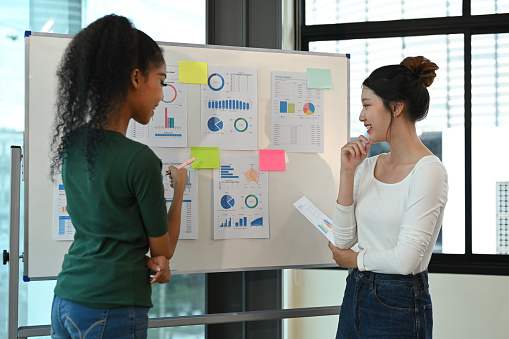 Two young creative women standing near flip chart with her colleague and discussing new marketing strategy together.