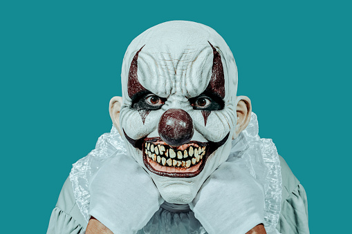a bald evil clown, wearing white gloves and a costume with a white ruff, staring at the observer with a creepy smile, leaning his head in his hands, on a blue background