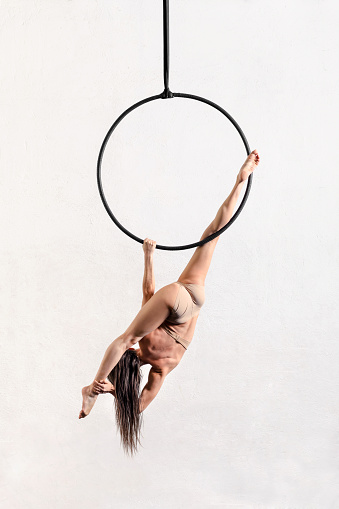 Full body back view of anonymous female aerialist performing split pose on aerial hoop against white background in light studio
