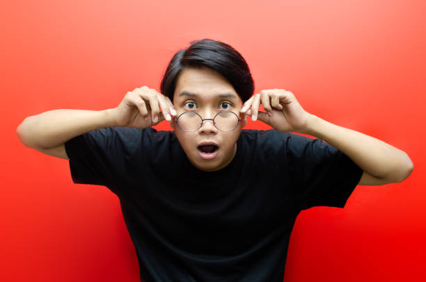 schockiertes gesicht eines asiatischen mannes, der die brille in schwarzem hemd auf rotem hintergrund senkt. - gasping stock-fotos und bilder