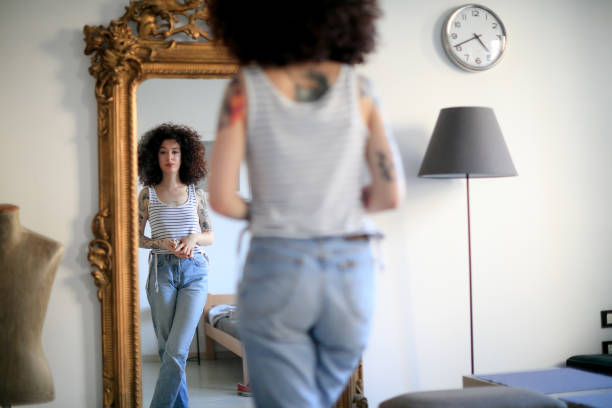 giovane donna tatuata con i capelli ricci che guarda il suo riflesso in specchio a figura intera a casa - thin curls foto e immagini stock