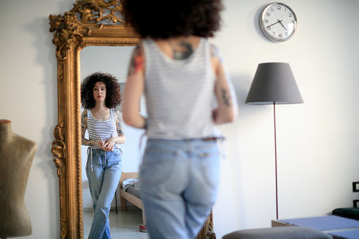 Young tattooed woman with curly hair standing cross-legged in front of ornate full length mirror and looking at her reflection
