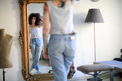 Young tattooed woman with hand in curly hair standing cross-legged in front of ornate full length mirror and looking at her reflection with attitude