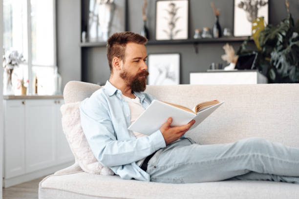 istock Young bearded handsome man reading book on couch at home 1427910039