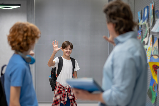 Smiling pleased cute schoolboy with the backpack waving his hand at his schoolmate and teacher