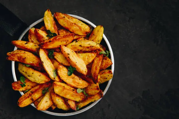 Photo of Roasted potatoes. Baked potato wedges in frying pan on dark stone background.