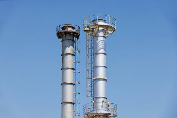 Two power plant metallic chimney towers with ladder and security fences on top, rising high against blue sky, environmental issues