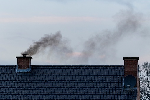A roof with two chimneys on it. In one of the chimneys there is a pipe mounted on it were a lot of black polluting smoke is coming out or rising up from, connected to the fireplace of the house.