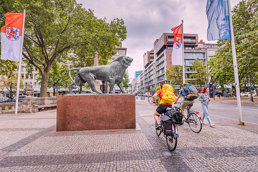 21 July 2022, Dusseldorf, Germany: Tourists cyclists at the statue of the lion - the symbol and coat of arms of Dusseldorf near the royal alley and canal