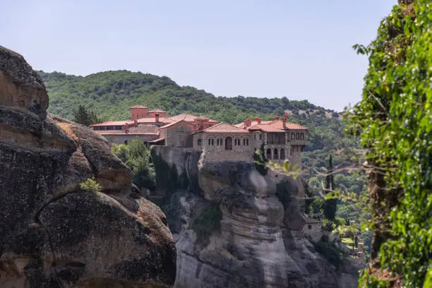 Photo of The Holy Varlaam Monastery is visible in crevice of neighboring rocks, it stands on monolith composed of sandstone and conglomerate, surrounded by lush vegetation, Meteora, Central Greece