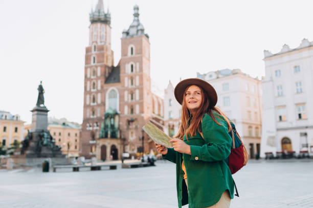 魅力的な若い女性観光客が新しい街を探索しています。クラクフのマーケット広場で紙の地図を持っている赤毛の少女。秋にヨーロッパを旅。セントメアリーズ大聖堂 - krakow people poland church ストックフォトと画像