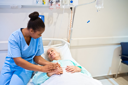Young female nurse consoling senior patient lying on gurney in hospital ward