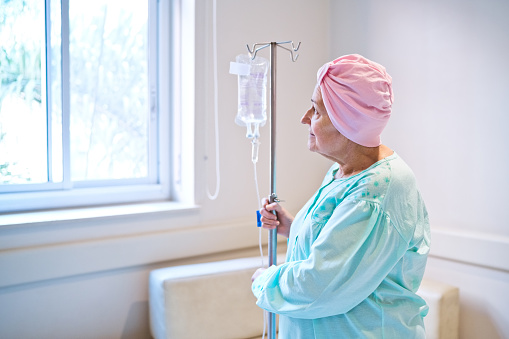 Senior woman wearing pink headscarf standing by IV drip in hospital ward