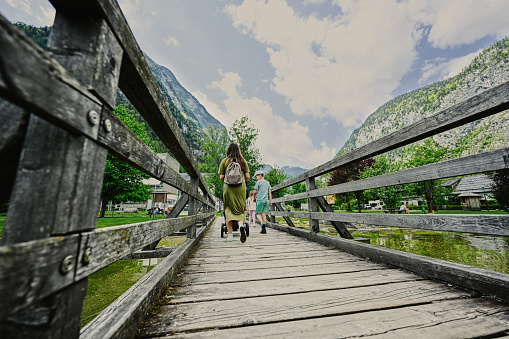 Back of mother with baby carriage and children walking on wooden bridge against Austrian alps in Hallstatt, Salzkammergut, Austria.