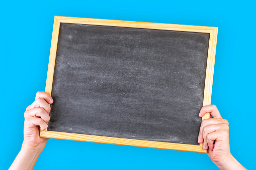 Front view of an unrecognizable white man hands holding a blackboard against a blue background. The chalkboard is clean so there is a useful copy space on it.