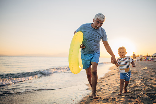 Grandfather walking on the beach with grandson and inflatable ring during sunset
