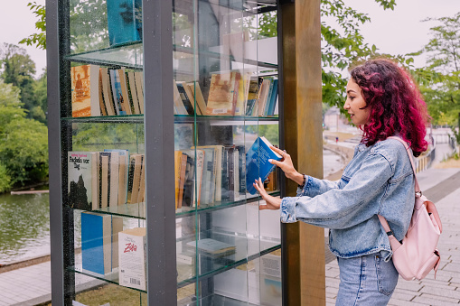 21 July 2022, Dusseldorf, Germany: Girl choosing books at free open public library at city park for book sharing among readers.