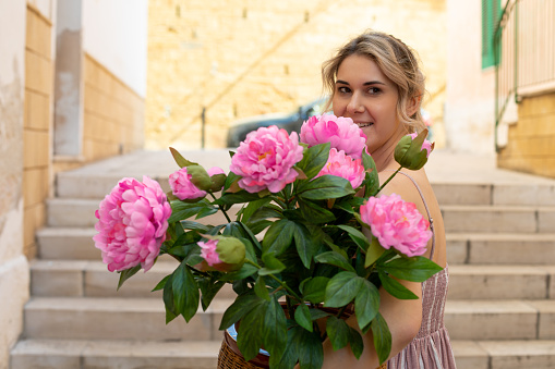 Portrait of young smiling woman with wavy fair hair wearing summer pink striped dress, standing on street near concrete stairs, holding brown pot with gorgeous artificial pink peonies flowers, posing.