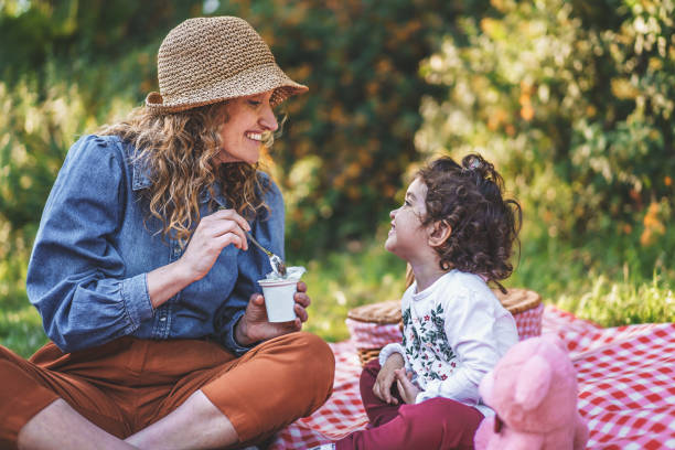 젊은 미혼모 숟가락은 야외 피크닉 중에 풀밭에 앉아있는 어린 딸에게 요구르트를 먹입니다. 가족, 야외 생활 및 분리 된 부모의 개념. - baby eating child mother 뉴스 사진 이미지