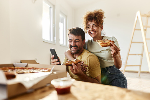 Young happy couple using mobile phone while eating pizza on a break from home renovation process. Copy space.