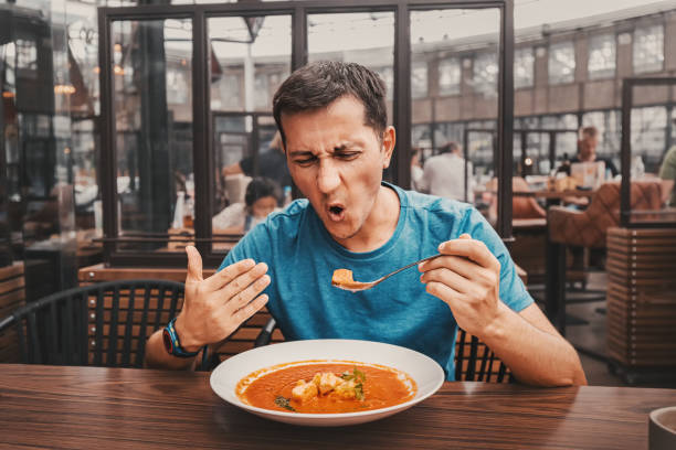 A man tries a spicy and hot red soup in a restaurant and reacts funny emotionally. Seasonings in the national cuisine and an unhealthy diet with overabundance of pepper stock photo