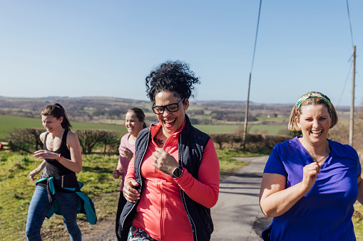 A group of mid adult women jogging uphill in County Durham, England with a scenic view behind them. They are wearing gym wear and smiling while they run.
