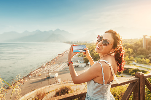 Travel blogger girl takes high-quality and vivid photos on the camera of his new expensive smartphone of the famous Konyaalti beach from a scenic viewpoint in Antalya, Turkey