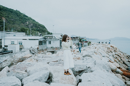 A portrait of a young happy woman wearing white dress with her camera strapped around her neck, standing on a big rock or stone in the rocky beach or seaside or coastline. taking a picture or photo with her camera as a female photographer or a traveler or tourist, while enjoying the calm water and wind in front of the sea and exploring a traditional Hong Kong fishing village and the mountains.