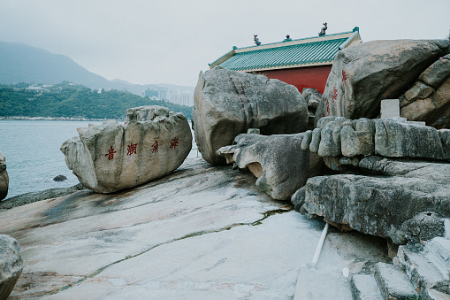 A view of a red ancient traditional old Chinese temple next to some big rocks or stones in the rocky beach or seaside or coastline, in front of the sea and a background of Hong Kong city with mountains and fog.