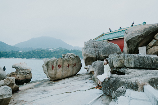 A portrait of a happy young Asian woman who is a traveler or tourist sitting on a big rock in front of a red ancient traditional old Chinese temple next to some big rocks or stones, with red Chinese wordings on them, in the rocky beach or seaside or coastline, in front of the sea and a background of Hong Kong city with mountains and fog, as she discovers and explores the fishing village or nature of Hong Kong.