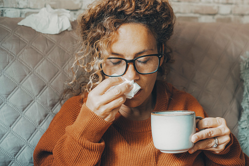 Mujer enferma en casa sonándose la nariz y cuidando de la enfermedad del virus de la influenza. Una mujer usa pañuelo de papel y bebe té de hierbas sola en casa. Concepto de resfriado gripal en temporada de invierno en interiores photo