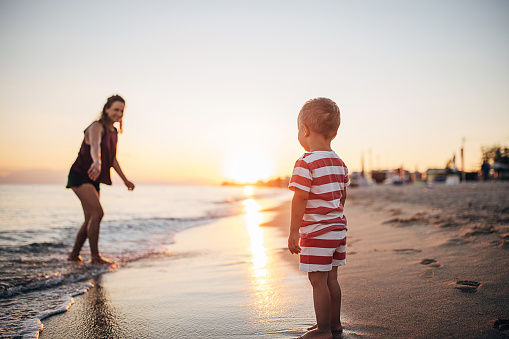 A child looking at his mother in the water on the beach in the sunset. She is looking at her child and calling him to follow her.