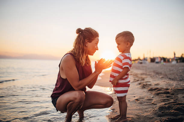A mother and her child on the beach in the sunset. She is crouching in the shallow water while he is standing.