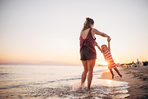 A mother and her son playing on the beach in the sunset. They are in the shallow water. She is holding and spinning him around.