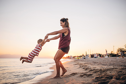 A mother and her child playing on the beach in the sunset. They are in the shallow water. She is holding and spinning him around.