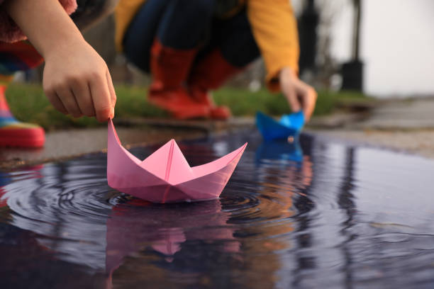 kleines mädchen und ihre mutter spielen mit papierbooten in der nähe von pfütze im freien, nahaufnahme - puddle rain boot water stock-fotos und bilder