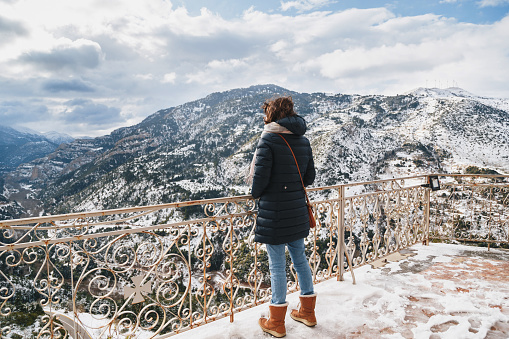 Back view of young woman standing at mountain top observation deck over Vouraikos gorge. Mega Spileon Monastery terrace with splendid snow-covered mountains view. Winter vacation. Greece.