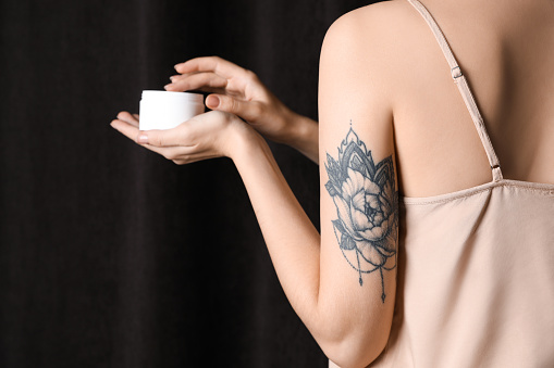 Woman with tattoo holding jar of cream against dark background, closeup