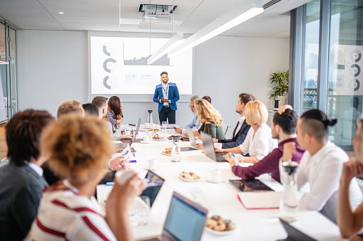 Wide angle view of businessmen and women sitting at board room conference table and watching smiling African male colleague give presentation.