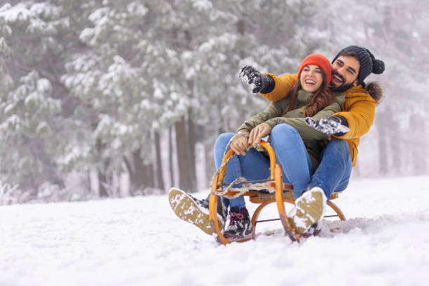 Couple sitting on sled and sliding down the hill while on winter vacation stock photo