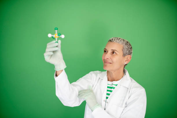 female scientist with white coat is holding model of molecule in her hand and is standing in front of green background