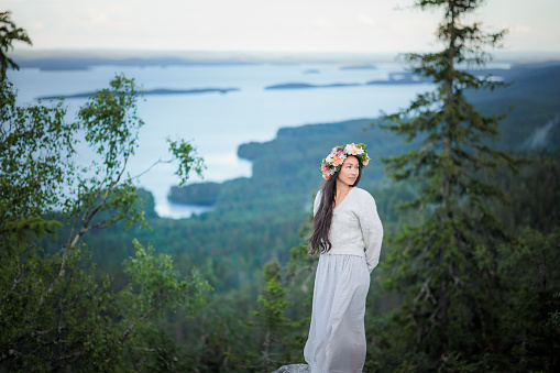 Caucasian-Chinese beautiful woman dressed in linen, fresh flower wreath, admiring the grand views of Koli National Park and Pielinen Lake in the mood of folklores